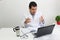 Latino medical doctor man with white coat works in his office with laptop, phone, coffee and glasses doing paperwork and video cal