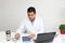 Latino medical doctor man with white coat works in his office with laptop, phone, coffee and glasses doing paperwork and video cal