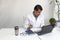 Latino medical doctor man with white coat works in his office with laptop, phone, coffee and glasses doing paperwork and video cal