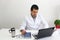 Latino medical doctor man with white coat works in his office with laptop, phone, coffee and glasses doing paperwork and video cal