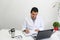 Latino medical doctor man with white coat works in his office with laptop, phone, coffee and glasses doing paperwork and video cal