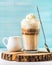 Latte macchiato with whipped cream, serving silver spoon and white pitcher on wooden round board over blue painted wall