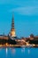 Latvia, Riga Evening Cityscape. View Of Illuminated St. Peter`s Church Tower Behind Daugava River Quay