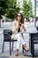 Laughing brown-haired woman is sitting in outdoor cafÃ© teracce in the street and using phone and text messaging