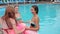 Laughing friends holding hands dance into swimming-pool with blue water, summer, joyful girls with pink inflatable rings