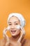 Laughing girl applying moisturizing cream on her face. Photo of young girl with flawless skin on orange background. Skin care and