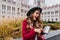 Laughing gorgeous woman in hat showing phone screen while exploring old part of city. Outdoor shot of smiling refined