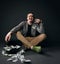 Laughing loudly middle-aged man in jacket, jeans and shoes sits on floor by upside down hat full of dollars cash