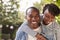 Laughing young black couple piggyback in garden, to camera