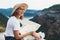 Laughing young blonde tourist holds in hands map of  landscape and looks views top of mountain landscape, cute smiling girl in hat