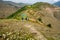 A layered cinematic shot of Himalayan Meadows with farmlands, and a traditional mud home in the background.with people going down