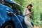 Leaning, using smartphone. Young woman is standing outdoors near electric automobile that is charging