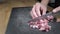 Left handed man cutting strips of bacon into cubes with big kitchen knife on black board on wooden kitchen table