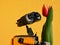 LEGO Wall-E robot from Pixar animated movie examining real orange tulip flower, bright yellow wall in background.