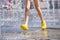Legs of a child in yellow rubber shoes running through the refreshing splashes of the city fountain on a hot summer day