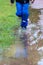 Legs of a young boy in blue waterproof raincoat and rain boots is jumping in deep puddles on a rainy day with happiness in splash