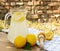 Lemonade in glass  jug / pitcher on wooden table with textile with lemon halves and wooden citrus pestle on brick wall background