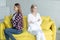 Lesbian females couple in casual clothes sitting back to back with crosed hands on yellow sofa after quarrel