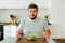 Lethargic young man sits in the kitchen in front of a bowl of greens with his hands on the table.