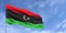 Libya flag on flagpole on blue sky background. Libyan flag fluttering in the wind against a background of sky with clouds. Place