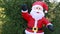 A life-size inflatable Santa Claus doll in a red suit stands swaying, waving against a background of green trees.
