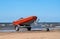 Lifeboat on trailer. The lifeboat is orange. The trailer with the boat is on the shore by the water. Sandy beach