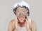 Lifestyle funny portrait of happy weird man on shower cap looking to himself in bathroom mirror with green cream on his face apply