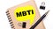 On a light background, a brown notebook, a black pen and paper clips, and a yellow card with the text MBTI
