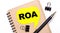 On a light background, a brown notebook, a black pen and paper clips, and a yellow card with the text ROA Return On Assets