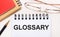 On a light background - glasses in gold frames, a pen, brown and red notepads and a white notebook with the text GLOSSARY.