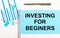 On a light background - light blue diagrams, paper clips and a sheet of paper with the text INVESTING FOR BEGINERS. View from