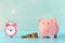 On a light blue background photographed pink piggy bank, next to her a small alarm clock and three stacks of coins