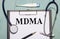 On a light blue wooden background, there is a paper labeled MDMA, a stethoscope, an electronic thermometer, and a pen. Medical