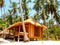 A light brown house with a straw roof stands on piles on a white sand beach