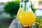 Light Bulb Glass Bottles with Fresh Orange Tropical Fruits Juice on Open Cafe Table at Sunset. Green Palm Trees Foliage Background