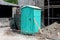 Light green portable ecological toilet left on construction site surrounded with sand and other construction material with old