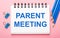 On a light pink background, light blue pencils, paper clips and a white notebook with the text PARENT MEETING