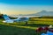 A light single-engine aircraft stands at a small rural airfield covered with green grass against a mountain landscape.