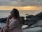 A light-skinned brown-haired girl in light beach clothes sits on the stones of the coastline of Karon beach in Phuket in Thailand