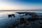 The light of a sunset casts golden highlights in pebbles and low rock formations on a beach under a purple and blue sky