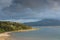 Light from sunset spotlights homes and a golden sandy beach along a calm bay under dramatic clouds