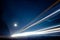 Light trails from vehicles traveling on rural road, surrounded by two rows of trees, late at night with moon on sky