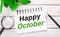 On a light wooden background, a magnifying glass, a pencil, a green plant and a white notebook with text HAPPY OCTOBER. Business
