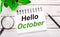 On a light wooden background, a magnifying glass, a pencil, a green plant and a white notebook with text HELLO OCTOBER. Business