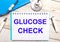 On a light wooden background there is a stethoscope, a blue notebook, a white pen and a sheet of paper with the text GLUCOSE CHECK