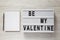 Lightbox with text `Be my Valentine`, blank notepad over white wooden surface. Valentine Day 14 February