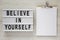 Lightbox with text `Believe in yourself`, clipboard with sheet of paper on a white wooden background, top view. From above, flat