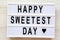 Lightbox with text `Happy Sweetest Day` word over white wooden background, top view. From above, flat lay, overhead