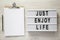 Lightbox with text `Just enjoy life`, clipboard with sheet of paper and pencil on a white wooden surface, top view
