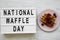 Lightbox with text `National Waffle Day` and traditional belgian waffle with raspberries on pink plate over white wooden surface